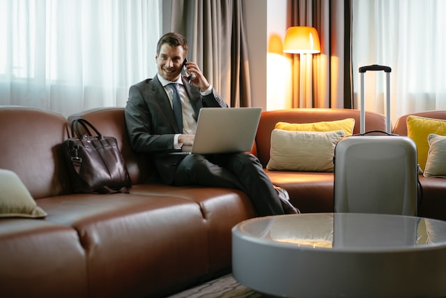 the businessman is sitting on the sofa with a laptop, there is a suitcase next to him